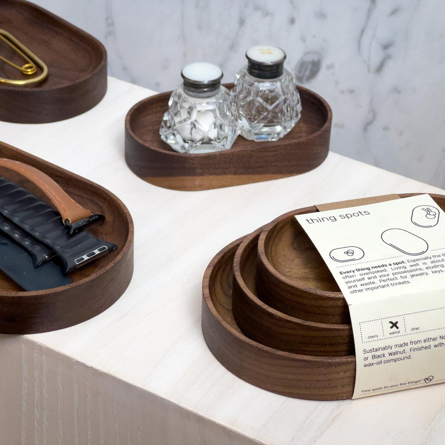The Thing Spots, by Biglow Woodcraft; a wooden tray set for small objects. Various sets of Thing spots with salt and pepper, and watch bracelets on a white table. Hardwood Walnut