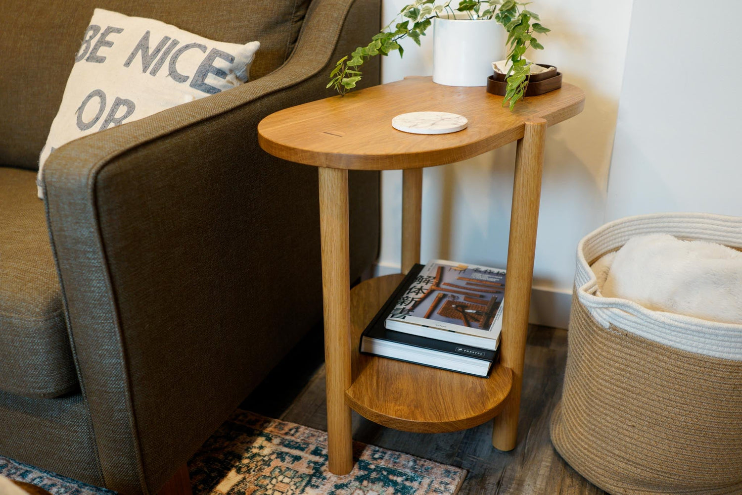 The Beside Table, by Biglow Woodcraft; a wooden table with a book, a plant and a coaster. Between a sofa and a basket. Hardwood White Oak