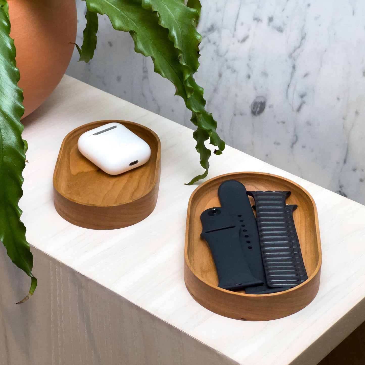 The Thing Spots, by Biglow Woodcraft; a wooden tray set for small objects. Two trays of Thing spots with watch bracelets, airpods and a plant on a white table. Hardwood White Oak