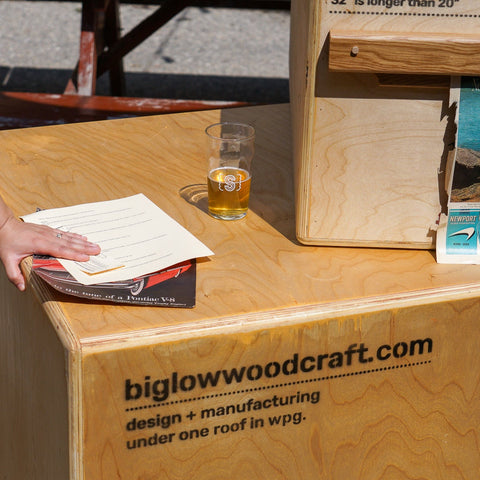A wooden display table with printed branding reading biglowwoodcraft.com, with a person partially visible on the left holding a drink and standing beside brochures and a wooden crate outdoors.