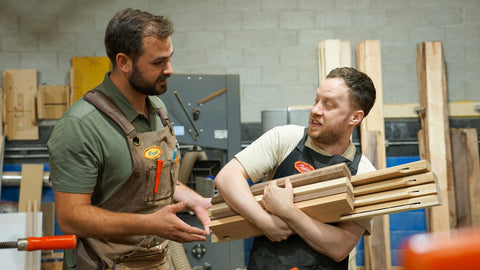 Two men in workshop aprons standing and talking, with one holding a stack of wooden Gravity Bars, a paper display product sold by Biglow Woodcraft. The setting is a woodworking shop with tools and lumber in the background.