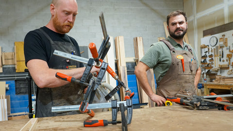 Two men in workshop aprons using orange-handled clamps on a wooden workbench in a woodworking shop.
