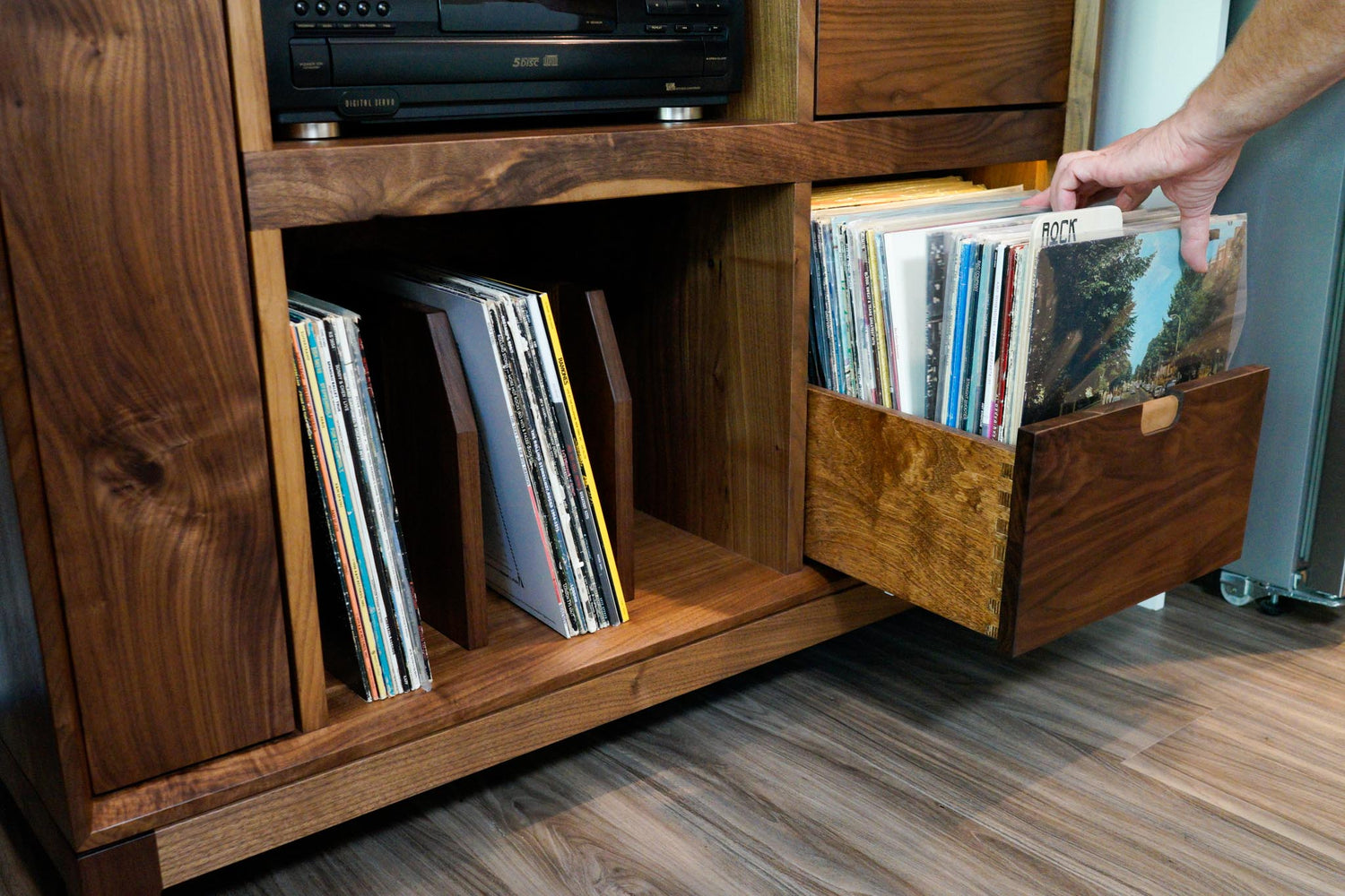 The lower half of a custom walnut record cabinet, showing a drawer being opened and sorted through. The drawer is full of records with custom genre dividers in between.