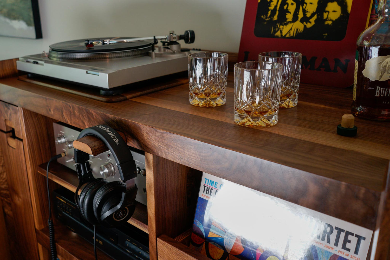 A detail shot of a custom walnut record cabinet, with a headphone rack poking out holding a pair of headphones. Scotch in 3 rocks glasses sit on the top, in front of a Doors record.