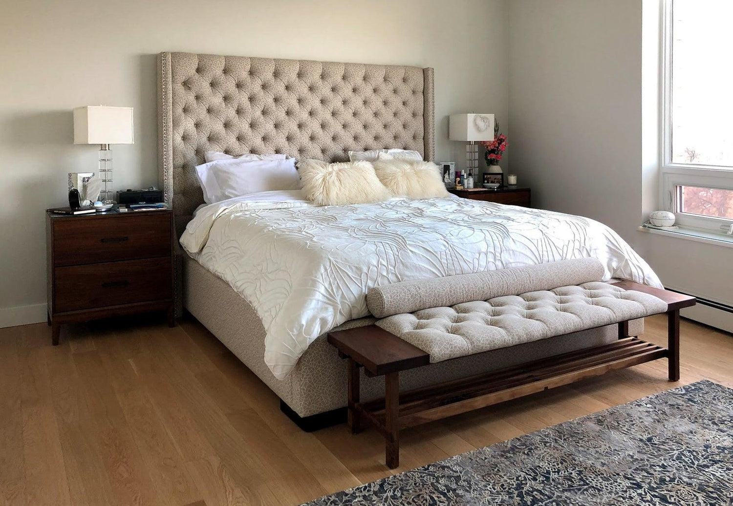 The custom walnut bedroom bench sitting at the foot of a bed. Its fabric matches that of the headboard, and its wood matches the side tables.