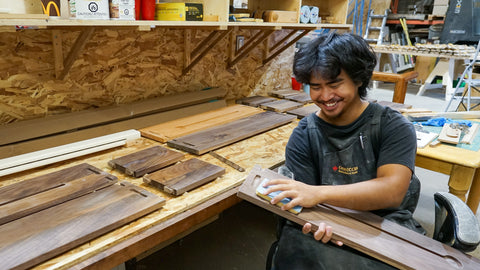 A smiling man wearing a black t-shirt and dark gray work apron sits in a woodworking workshop, holding and sanding a walnut panel. The background features a wooden workbench, shelves with various tools, and wood panels being made into the Biglow Woodcraft Book Stop.