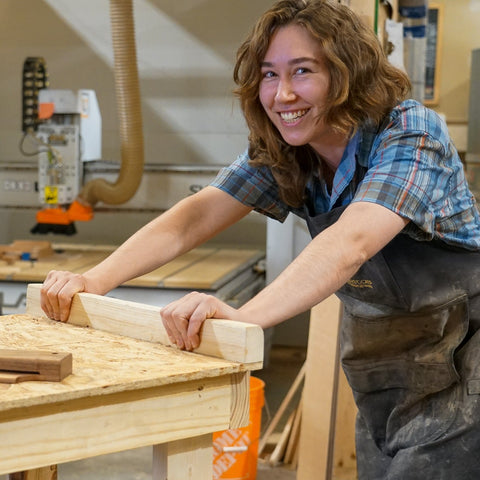 A smiling woman in a woodworking shop leans over a workbench holding a piece of wood, with woodworking equipment and materials visible in the background.