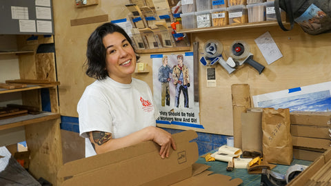 A smiling woman with dark hair stands in a workshop, wearing a white t-shirt with a red logo and holding a cardboard box. The background is filled with shelves, tools, and packaging materials.