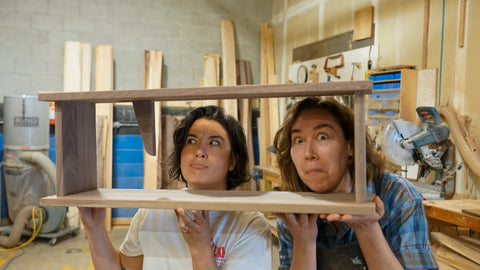Two people in a woodworking workshop holding up a wooden book shelf called the Book Stop, with wood scraps and tools visible in the background.