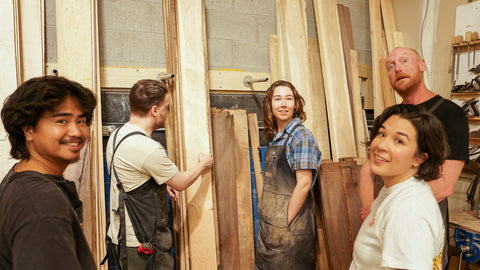 Five people standing in a wood workshop, with tall vertical lumber stacks behind them. The group includes three men and two women, some wearing work aprons, and one man in the foreground looking toward the camera.