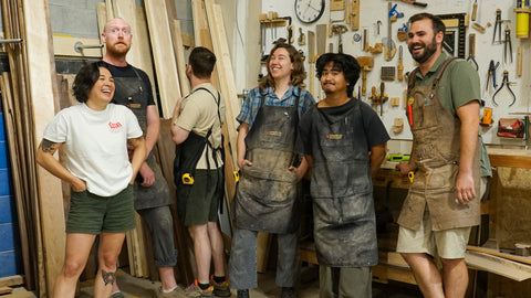 Six people wearing work aprons stand together in a woodworking workshop filled with fine woodworking tools and hardwood.