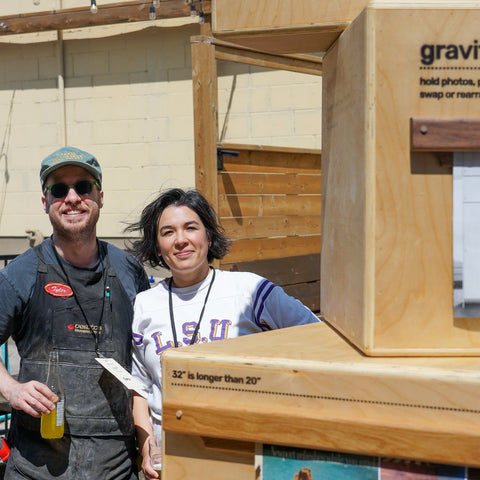 Two people standing outdoors next to a large wooden display labeled 'gravity bar' with printed instructions and a photo insert, in front of a beige building.