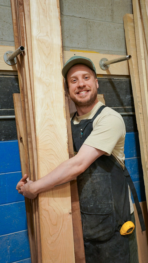 A smiling man wearing a green cap, beige t-shirt, and dark gray work apron leans against a stack of wooden boards in a workshop setting.
