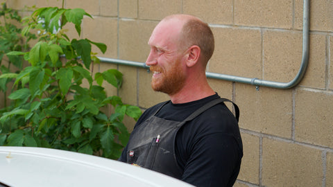 A man with a shaved head and beard, wearing a black t-shirt and dark gray utility apron, smiling while sitting outdoors next to a white vehicle in front of a beige brick wall.