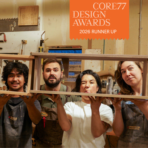 Four people in a woodworking workshop holding a wooden book shelf, with an orange 'CORE77 DESIGN AWARDS 2026 RUNNER UP' badge in the top right corner.