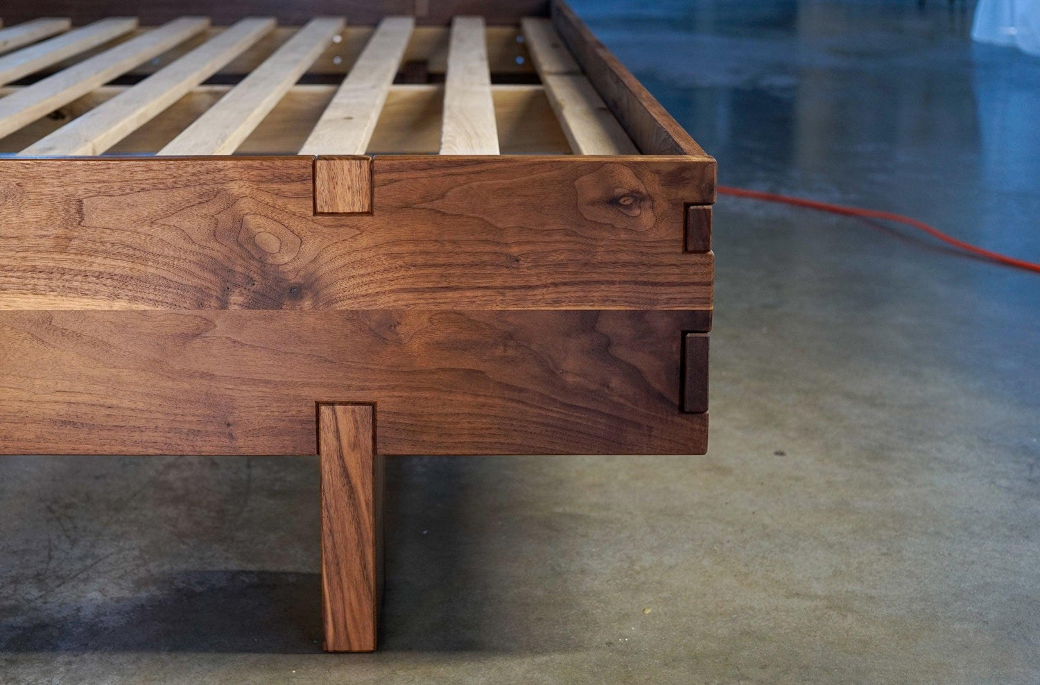 The foot of a custom walnut bed frame, showing a fancy detail where the leg joins the frame, and the oversized finger joints on the corner of the frame itself.